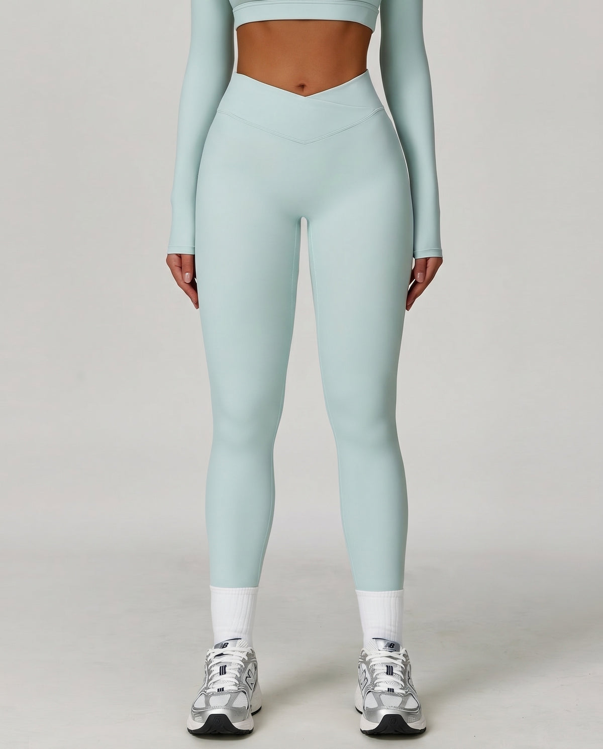Front view of high waist crossover sculpt compression leggings in light blue, featuring a V-shaped crossover waistband and full-length squat-proof fit paired with a matching long sleeve crop top and white sneakers.