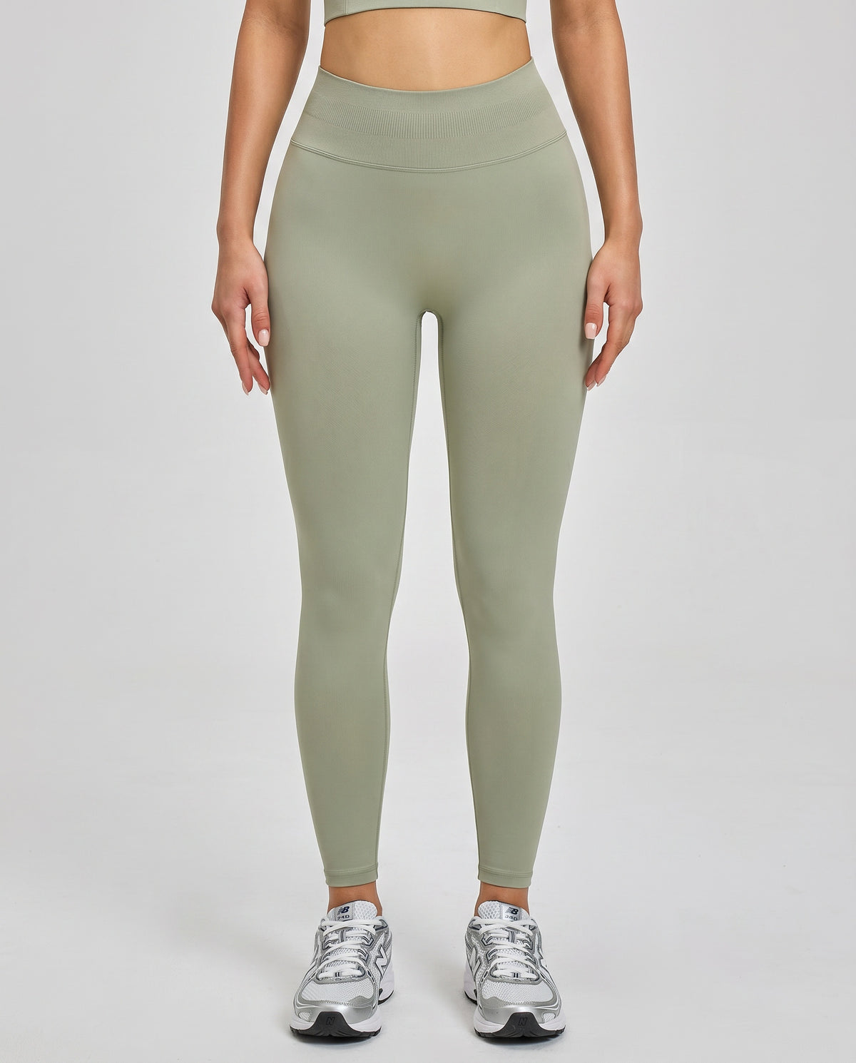 Front view of seamless high waist compression leggings in sage green with wide sculpting waistband and ankle-length fit, paired with sneakers.