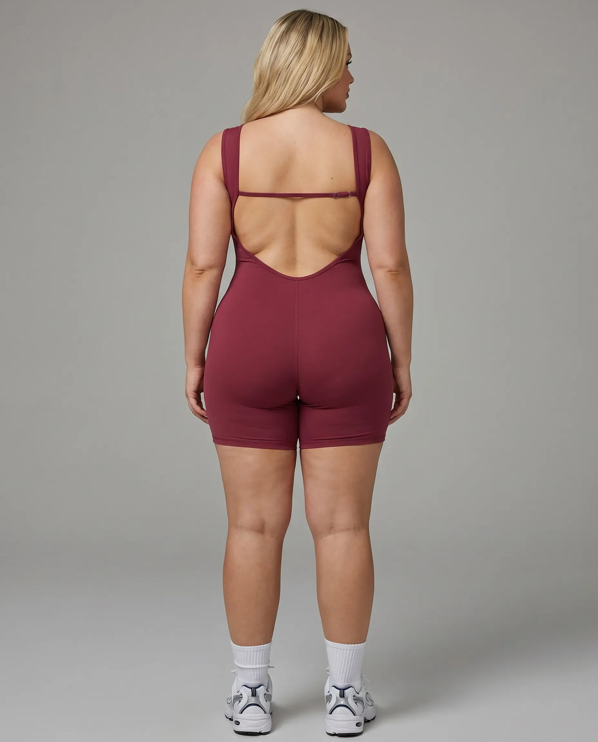 Female model wearing red backless sculpting bodysuit, rear view showing open back detail in studio.