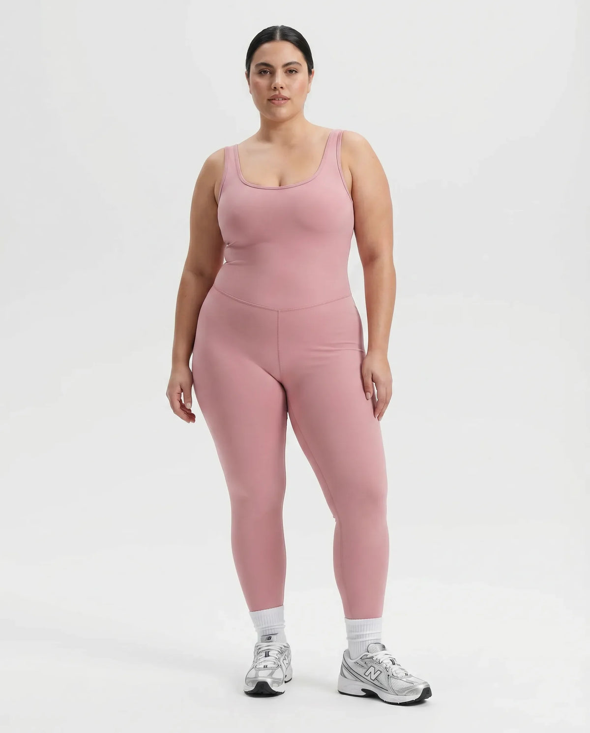 Female model standing front-on in a studio wearing pink sculpting backless bodysuit with white sneakers.
