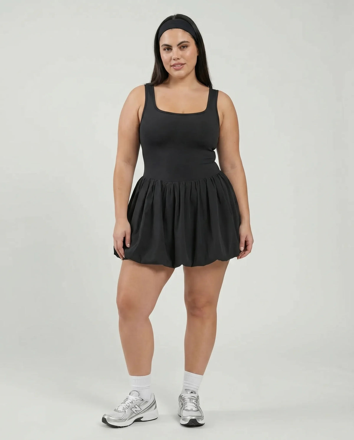 Model facing forward wearing black balloon activewear dress with white sneakers in a studio setting.