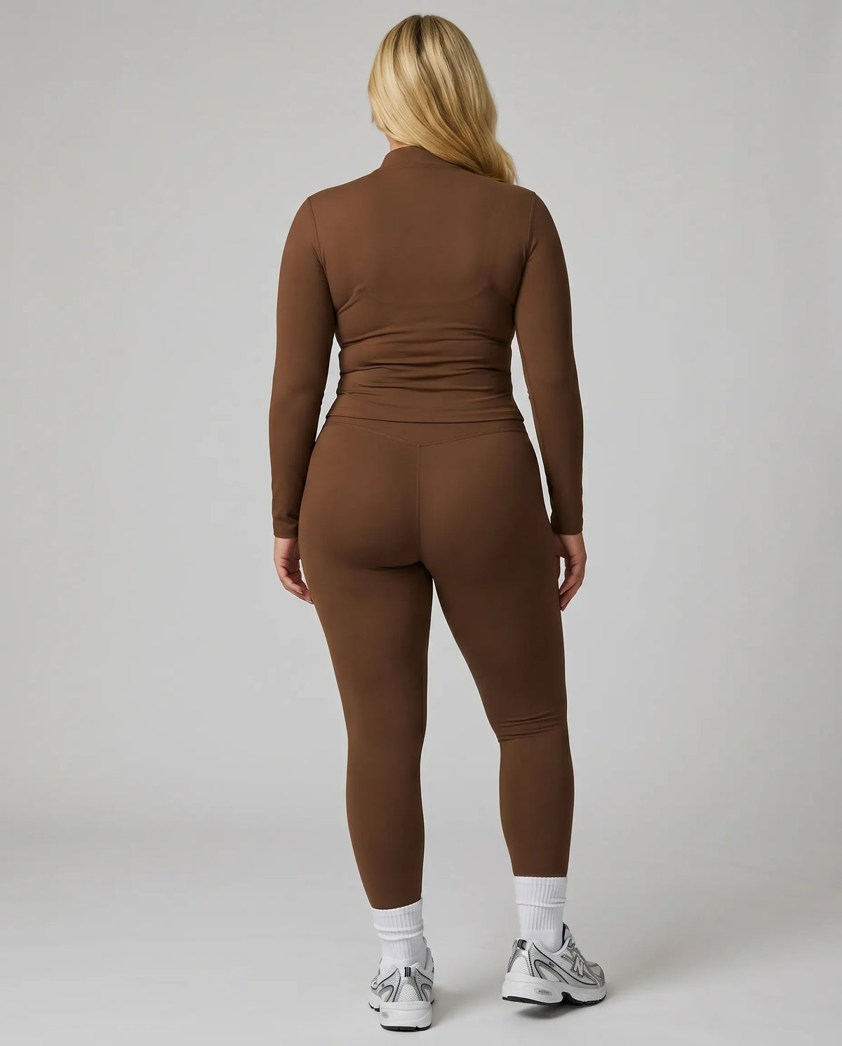 Female model facing away in studio showing the back fit of the brown ribbed zip jacket and matching leggings.