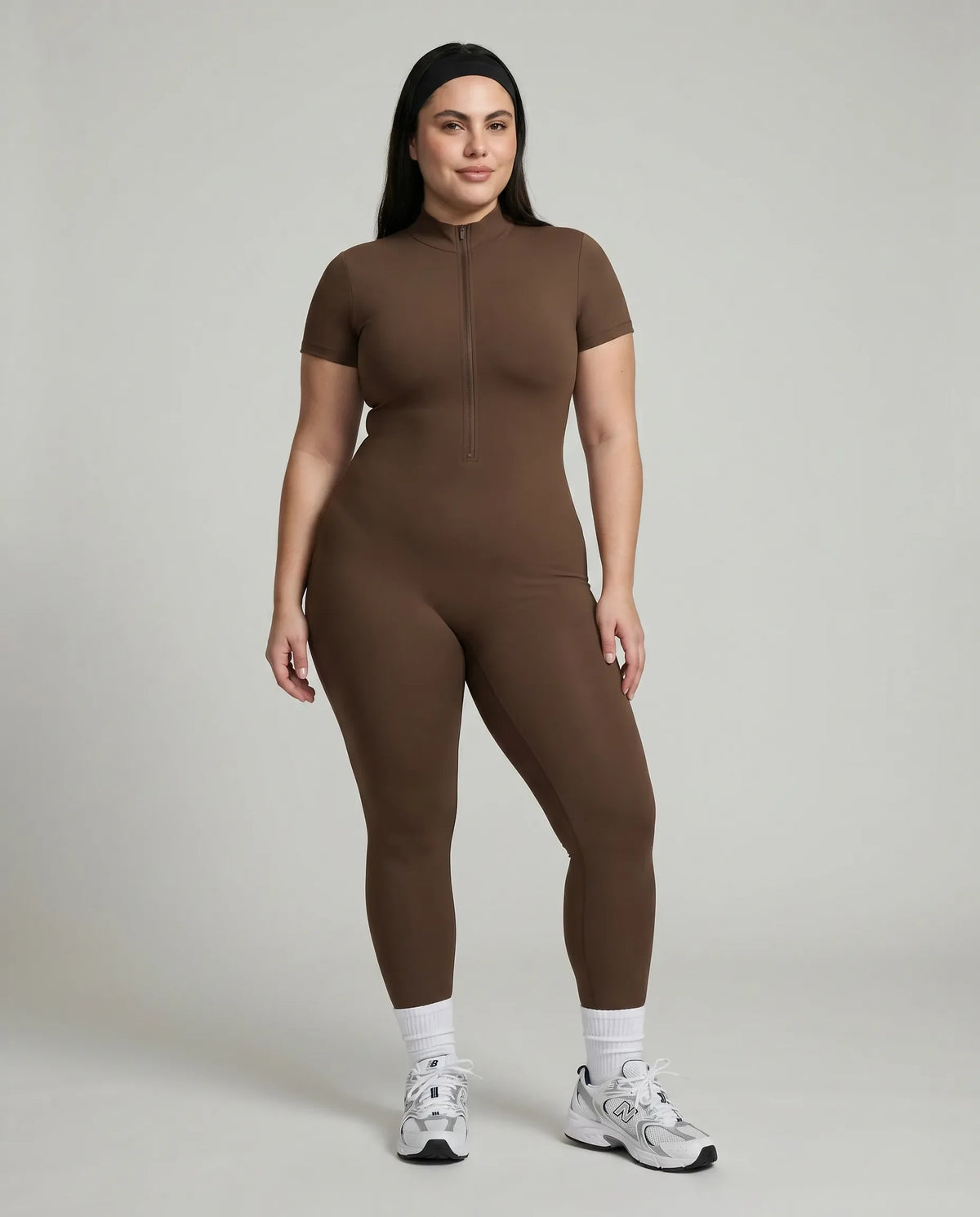 Female model standing front-on in studio wearing black short-sleeve compression jumpsuit with half-zip and white sneakers.