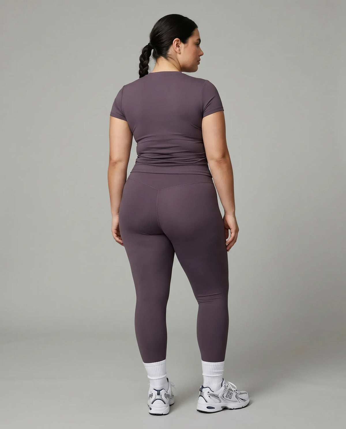 Model standing with back to camera in studio wearing purple fitted t-shirt top showing rear fit and silhouette.