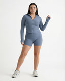 Woman wearing a fitted activewear set with high waist shorts and a zip-up jacket in a clean studio setting.