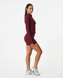 Woman wearing a fitted activewear set with high waist shorts and a zip-up jacket in a clean studio setting.