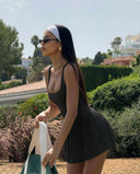 Model wearing black pleated tennis dress with white sneakers, standing in a front-facing studio pose.