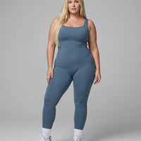 Model standing front-on in a blue backless flare bodysuit in a studio setting with arms relaxed.