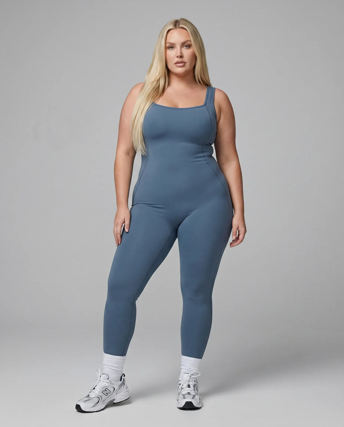 Model standing front-on in a blue backless flare bodysuit in a studio setting with arms relaxed.