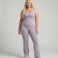 Model standing front-on in studio wearing purple flared leg backless bodysuit with white sneakers.