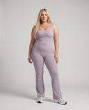 Model standing front-on in studio wearing purple flared leg backless bodysuit with white sneakers.