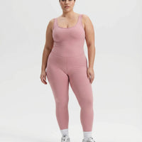 Female model standing front-on in a studio wearing pink sculpting backless bodysuit with white sneakers.
