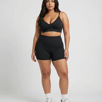 Model standing front-on in a studio wearing a black sculpting bra top paired with black bike shorts and sneakers.