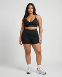 Model standing front-on in a studio wearing a black sculpting bra top paired with black bike shorts and sneakers.