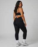 Model standing with her back to camera in a studio wearing a black V-neck bra top showing rear strap layout.