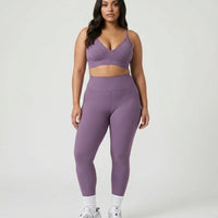 Model standing front-on in a studio wearing purple sculpting bra top and matching high waist leggings with white sneakers.