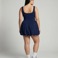 Female model photographed from behind in a studio wearing the badge blue balloon hem dress showing the back bodice and skirt volume.
