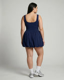 Female model photographed from behind in a studio wearing the badge blue balloon hem dress showing the back bodice and skirt volume.