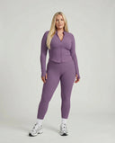 Model standing front-on in studio wearing purple sculpting ribbed zip jacket and matching high waist leggings.