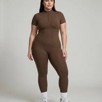 Female model standing front-on in studio wearing black short-sleeve compression jumpsuit with half-zip and white sneakers.