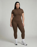 Female model standing front-on in studio wearing black short-sleeve compression jumpsuit with half-zip and white sneakers.