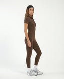 Woman in black zip-front jumpsuit mid-movement during a reformer pilates session in natural light studio.