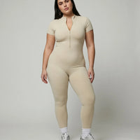 Model standing front-on in studio wearing yellow zip-front compression jumpsuit with white sneakers.