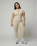 Model standing front-on in studio wearing yellow zip-front compression jumpsuit with white sneakers.