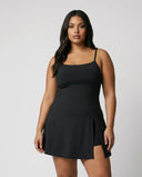Model wearing black padel dress with built-in shorts and white sneakers standing in studio front view showing sculpting fit and side slit detail.