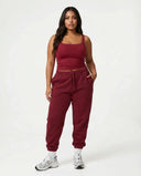 Female model standing front-on in studio wearing red sleeveless crop tank top with matching red joggers and white sneakers.
