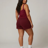 Female model standing with back to camera in studio wearing red sleeveless tank top showing back fit and strap detail.
