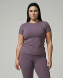 Model standing front-on in studio wearing purple fitted t-shirt top and matching leggings with white sneakers.