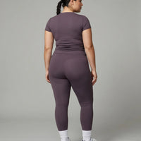 Model standing with back to camera in studio wearing purple fitted t-shirt top showing rear fit and silhouette.
