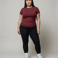Female model standing front-on in studio wearing red fitted crew neck tee with black leggings and sneakers.