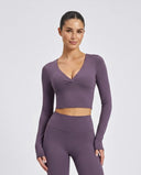 Woman wearing a purple two-piece sculpting activewear set with long sleeve V-neck crop top and high waist compression leggings, styled with white sneakers against a light grey background.
