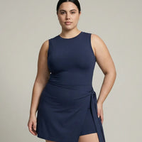 Model wearing blue sleeveless active dress with wrap skirt detail, front view in studio with white sneakers and headband.