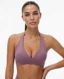 Woman wearing a purple deep V halter sports bra with sculpting compression fit and crossover band detail, styled with matching activewear bottoms against a white background.