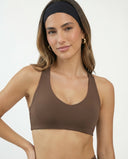 Model wearing a mocha scoop-neck seamless sports bra with wide racerback straps and compression fit, styled with a black headband and gold hoop earrings.
